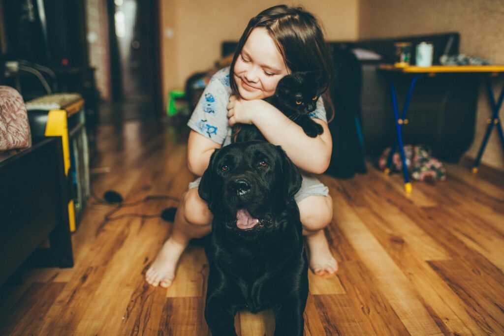 A smiling child hugs a Labrador and cat on a wooden floor, conveying warmth and happiness.